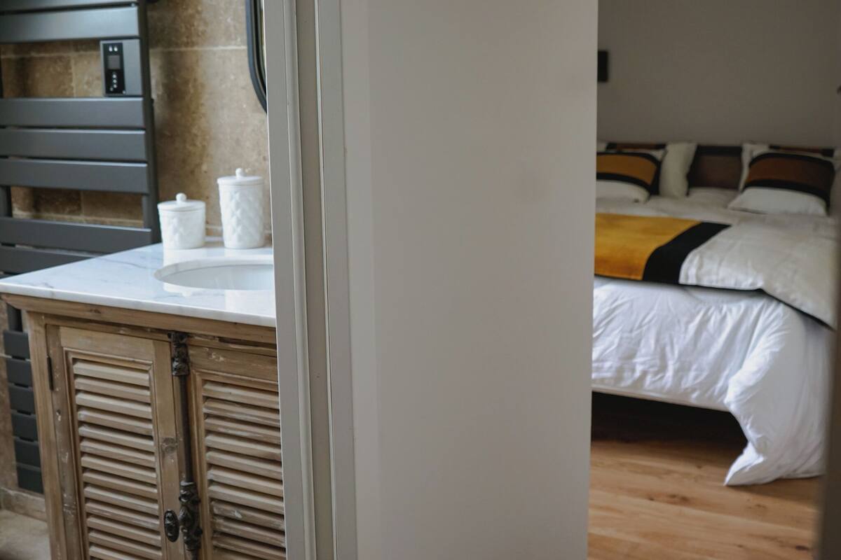 A hotel room with a wooden floor, a bed with white bedding and a yellow throw, a white sink with two containers, and a wooden cabinet with louvered doors.