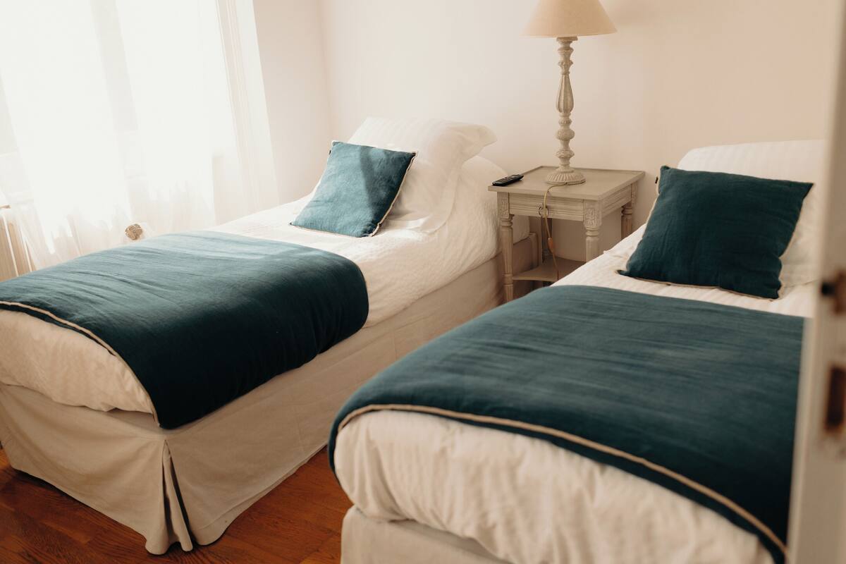 Two twin beds with teal bedding, a bedside table with a lamp, and a window with sheer curtains.