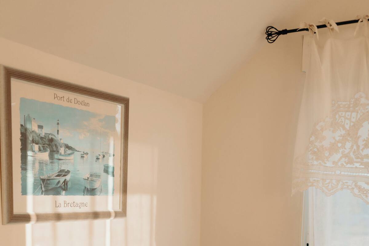 A beige sofa with a remote control on a wooden table, a framed picture of a coastal scene, and a sheer white curtain.