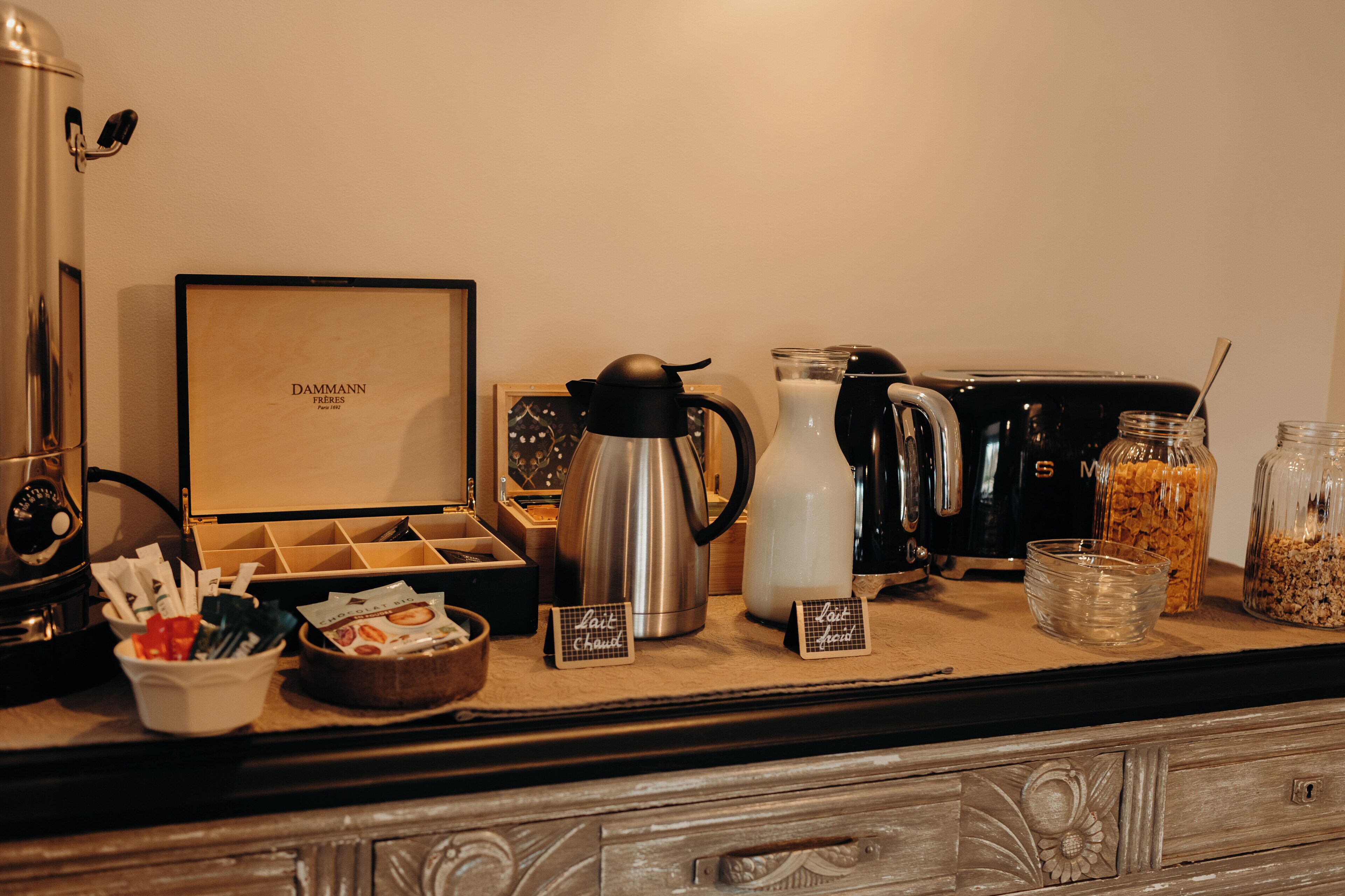 Un espace dédié au café, équipé de divers appareils pour préparer le café, notamment une machine à café, un mousseur à lait et une cafetière. On y trouve également des récipients contenant du café en poudre et du sucre, ainsi qu'un bol rempli de dosettes de café.