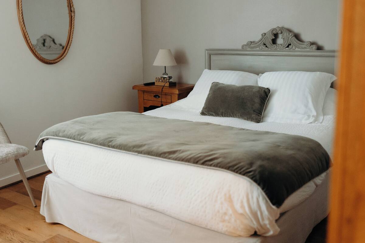 A neatly made bed with a grey and white duvet, a wooden nightstand with a lamp, and a chair in a room with light-colored walls and wooden flooring.