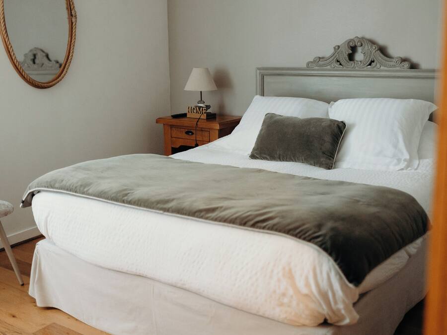A neatly made bed with a grey and white duvet, a wooden nightstand with a lamp, and a chair in a room with light-colored walls and wooden flooring.