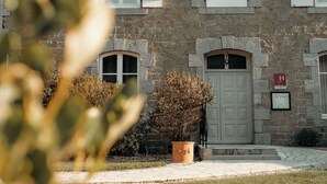 A stone building with a large central door, multiple windows with shutters, and a small garden area with a potted plant. - Hôtel Le Chatellier – Piscine chauffée d’avril à octobre (Cancale)