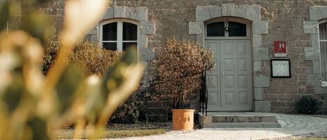 A stone building with a large central door, multiple windows with shutters, and a small garden area with a potted plant.