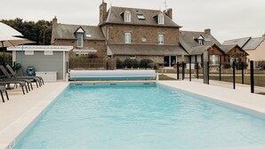 A rectangular swimming pool with a tiled deck, surrounded by a fence, and a building with a stone facade in the background. - Hôtel Le Chatellier – Piscine chauffée d’avril à octobre (Cancale)