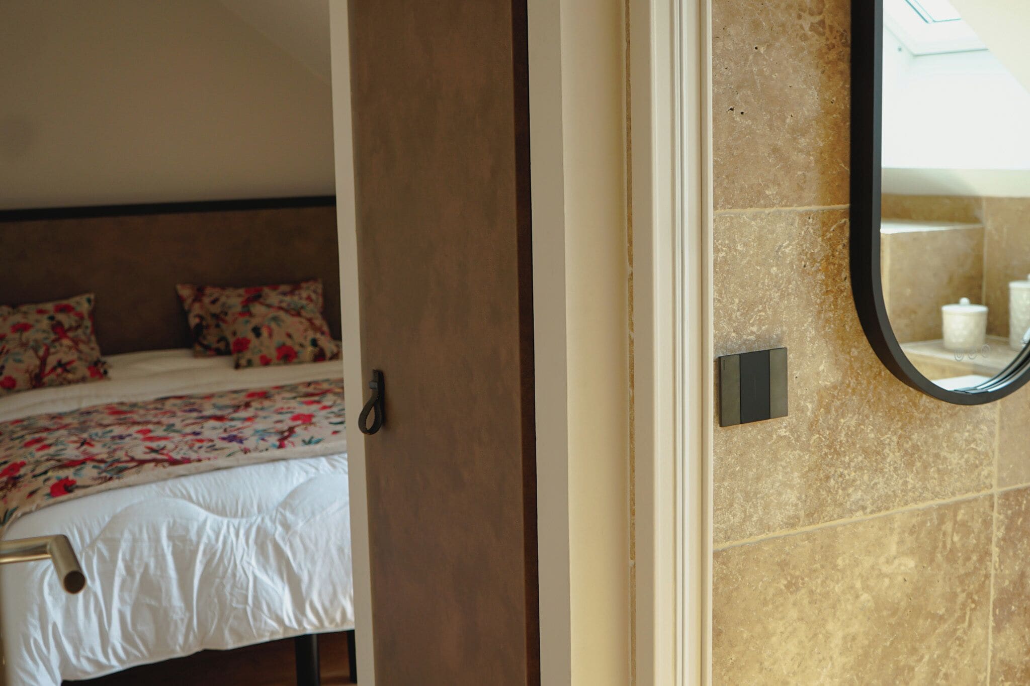 Een slaapkamer met een bed, bloemenbeddengoed, een hoofdbord en een badkamer met een betegelde wand en een spiegel.