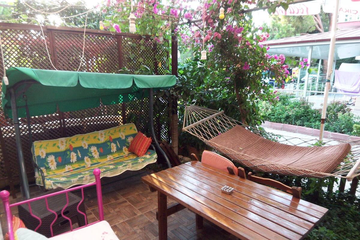 A wooden table and a hammock in a garden area with a wooden bench and a cushioned seat with a floral pattern.