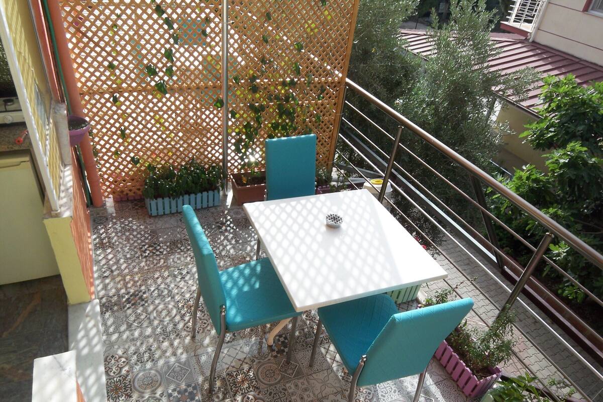 A balcony with a white table and blue chairs, surrounded by potted plants and a decorative fence.