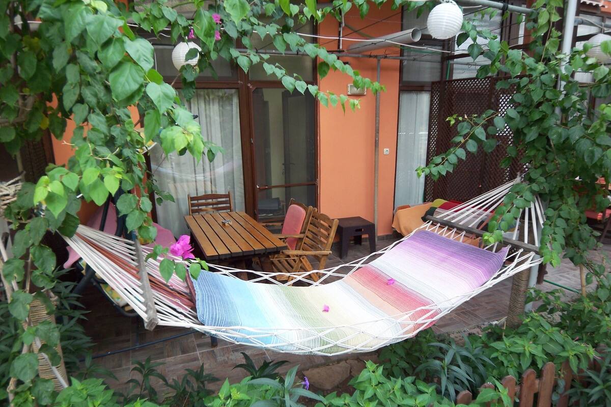 A hammock in a garden with a wooden table and chairs, surrounded by greenery.