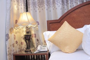 Una cama bien hecha con cabecera de madera, una lámpara de noche, un teléfono y una cortina con estampado floral.