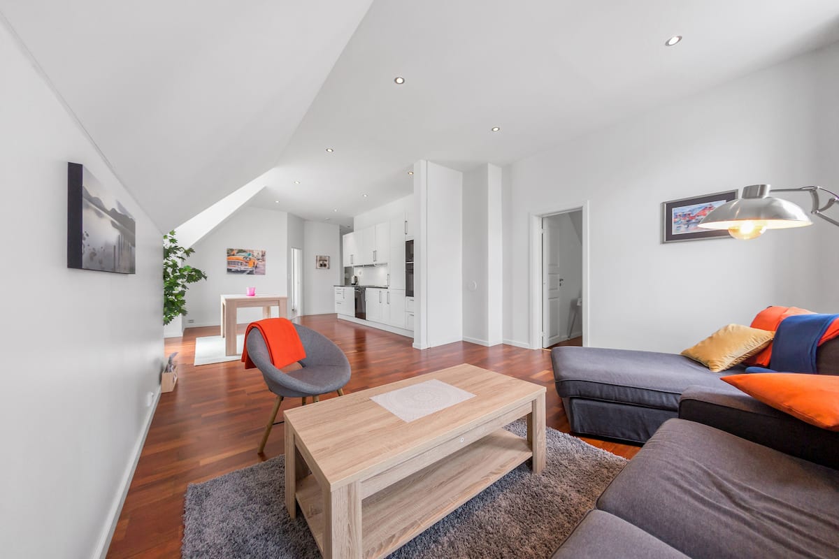Een moderne woonkamer met een houten salontafel, een grijze bank met oranje kussens en een grijze fauteuil.