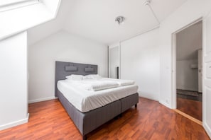 A bedroom with a bed, wooden flooring, a sloped ceiling, and a door leading to another room.