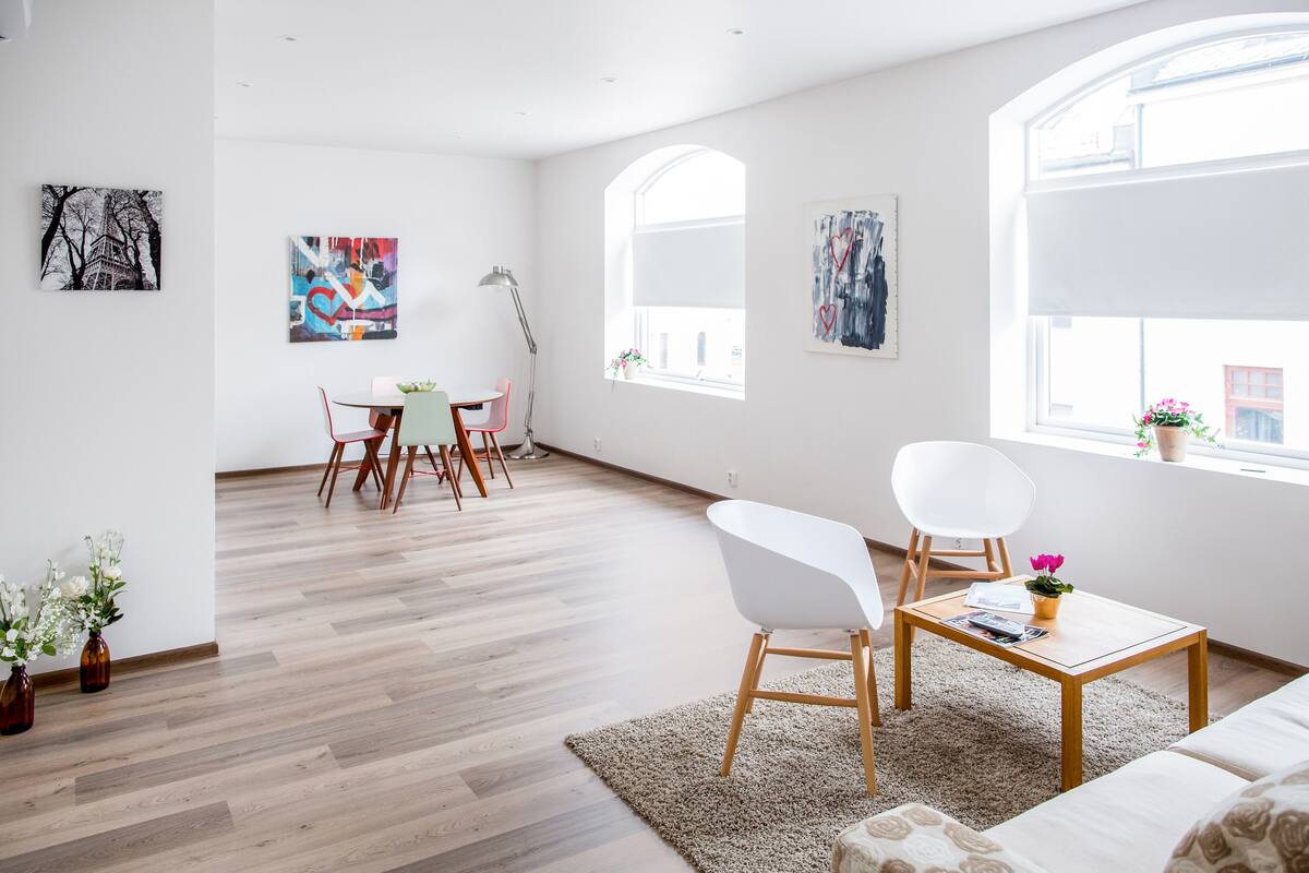 Een ruime woonkamer met een houten vloer, een witte bank, een salontafel en een eethoek met stoelen en een tafel.