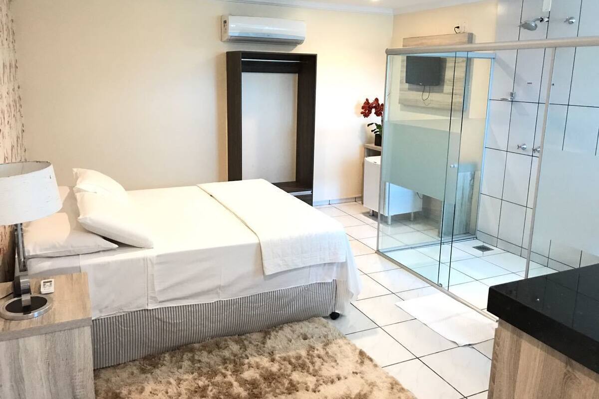 A modern hotel room with a bed, a desk, a mirror, a shower area, and a small seating area.
