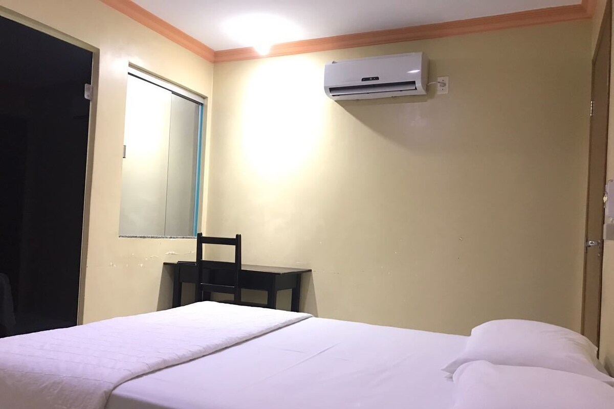 A hotel room with a bed, a chair, a desk, and an air conditioning unit.