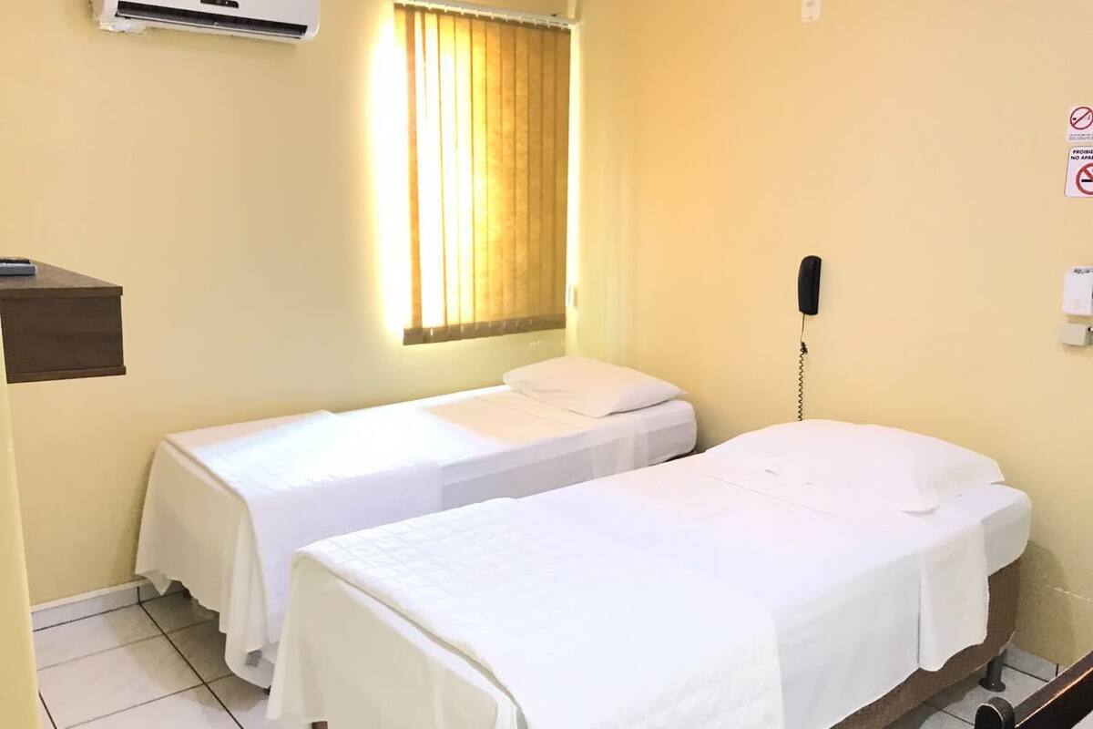 A hotel room with two single beds, a bedside telephone, an air conditioning unit, and a window with curtains.