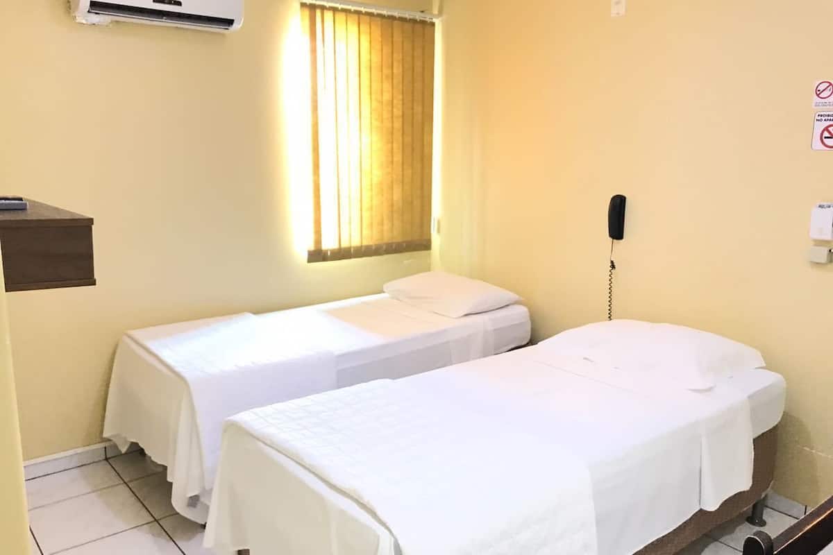A hotel room with two single beds, a bedside telephone, a window with curtains, an air conditioning unit, and a small shelf.