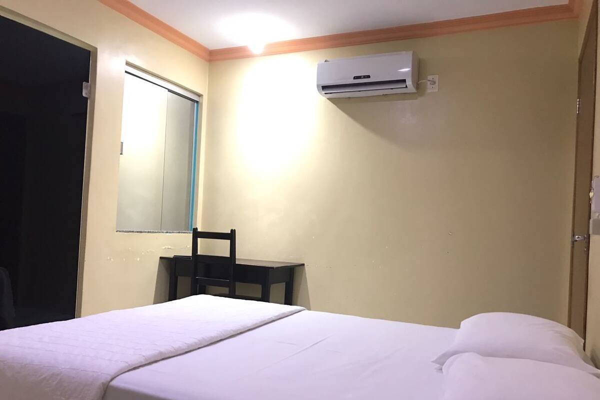 A hotel room with a bed, a chair, a desk, and an air conditioning unit.