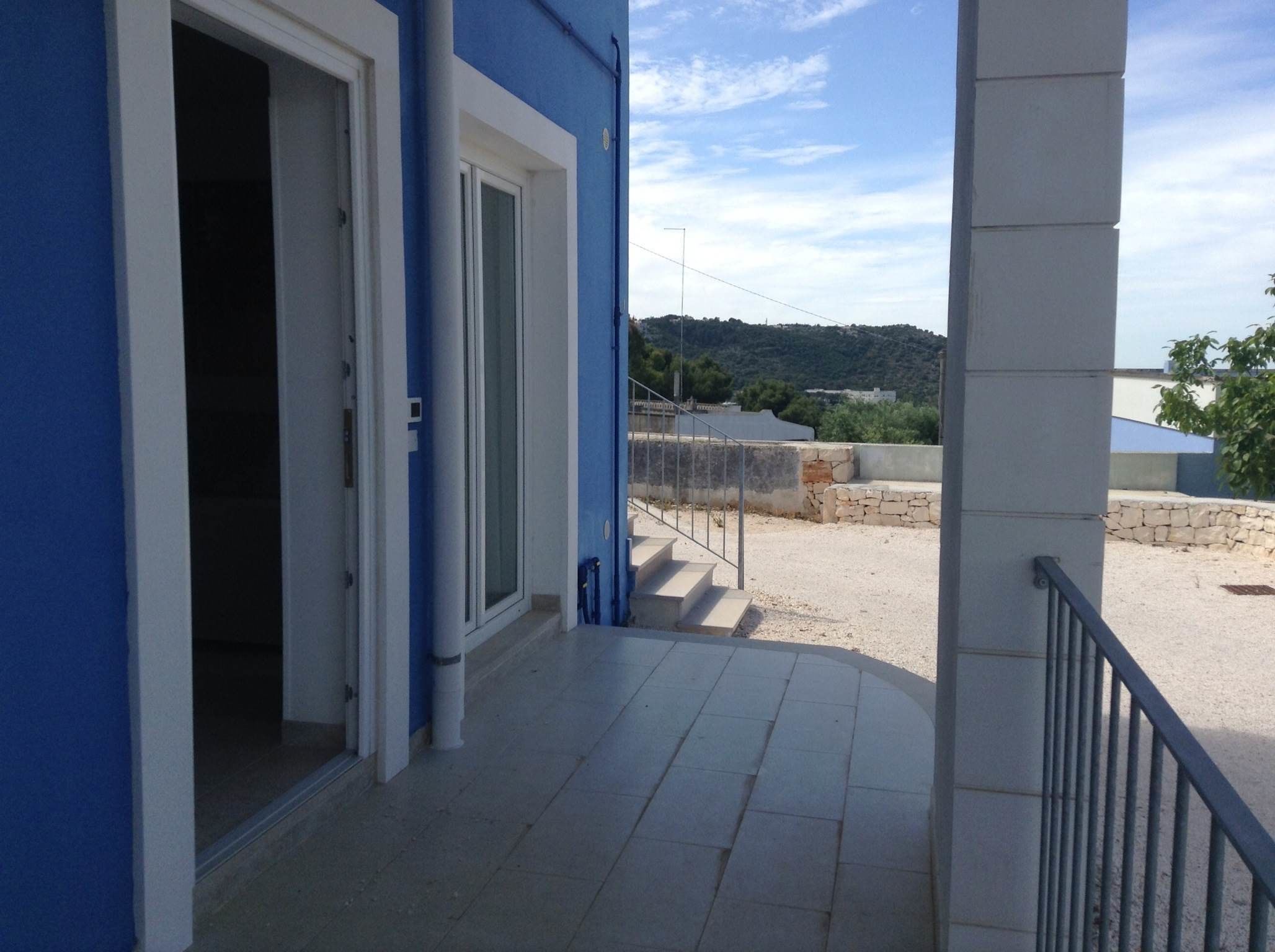 Un edificio de fachada azul, un espacio al aire libre techado con suelo de grava y una vista a una colina en el fondo.