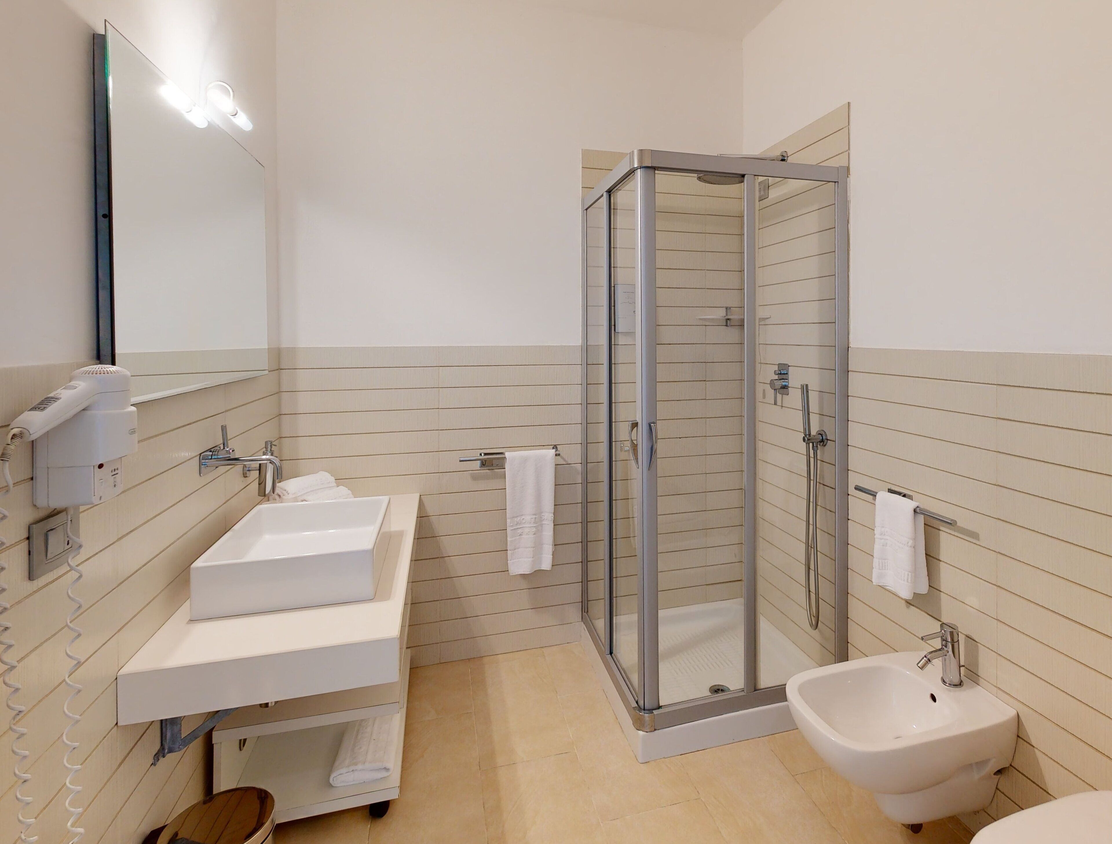 A modern bathroom with a glass-enclosed shower, a white sink, a bidet, and a mirror with lights.