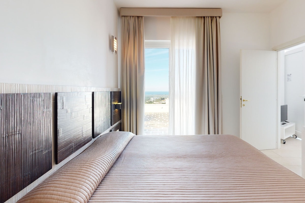 A hotel room with a large bed, a desk with a computer, and a view of the city through the window.