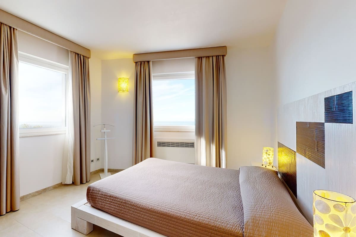 A hotel room with a bed, curtains, a bedside table, a lamp, and a view of the sea.
