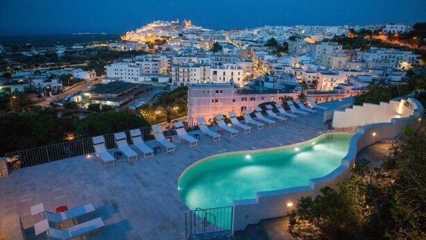 Zona de piscina con sillas de descanso y vista a un pueblo costero al atardecer.
