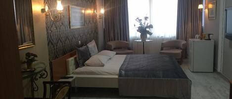 A hotel room with a bed, two armchairs, a small table, a refrigerator, and a mirror.