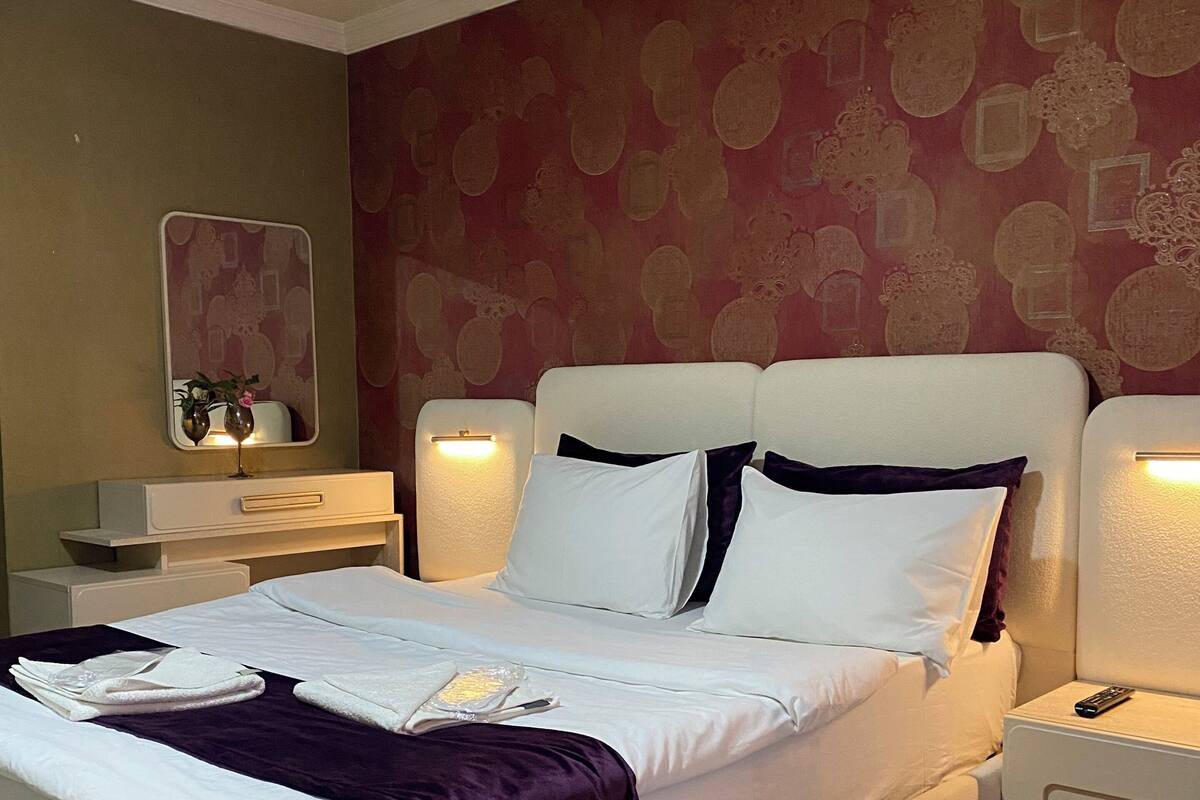 Una cama bien hecha con sábanas blancas y una manta morada, una mesita de noche con control remoto y una pared con papel tapiz estampado.