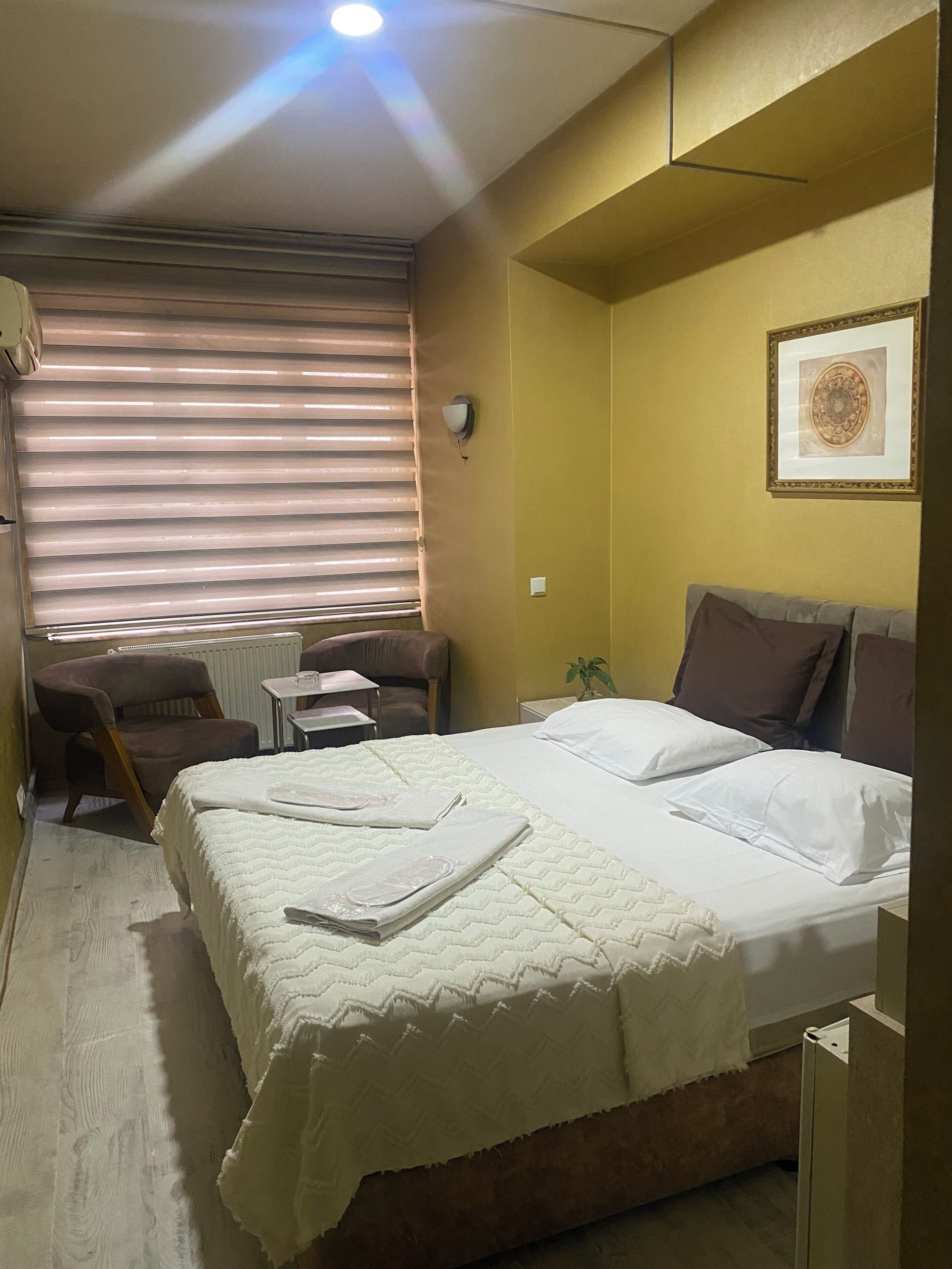 A hotel room with a bed, a small table, a chair, a window with blinds, and a wall-mounted light fixture.