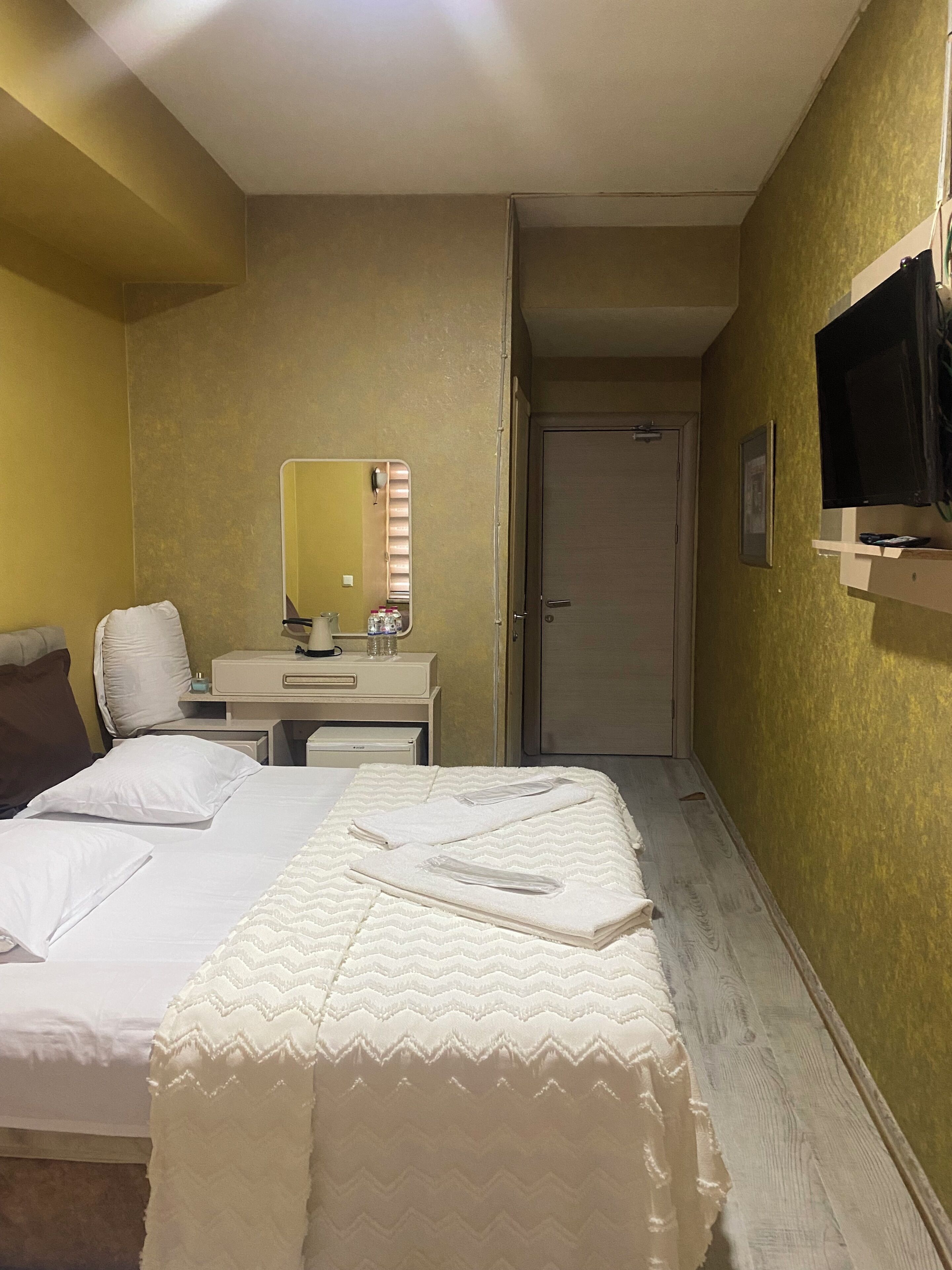A hotel room with a bed, a desk with a mirror, a television, and a door.