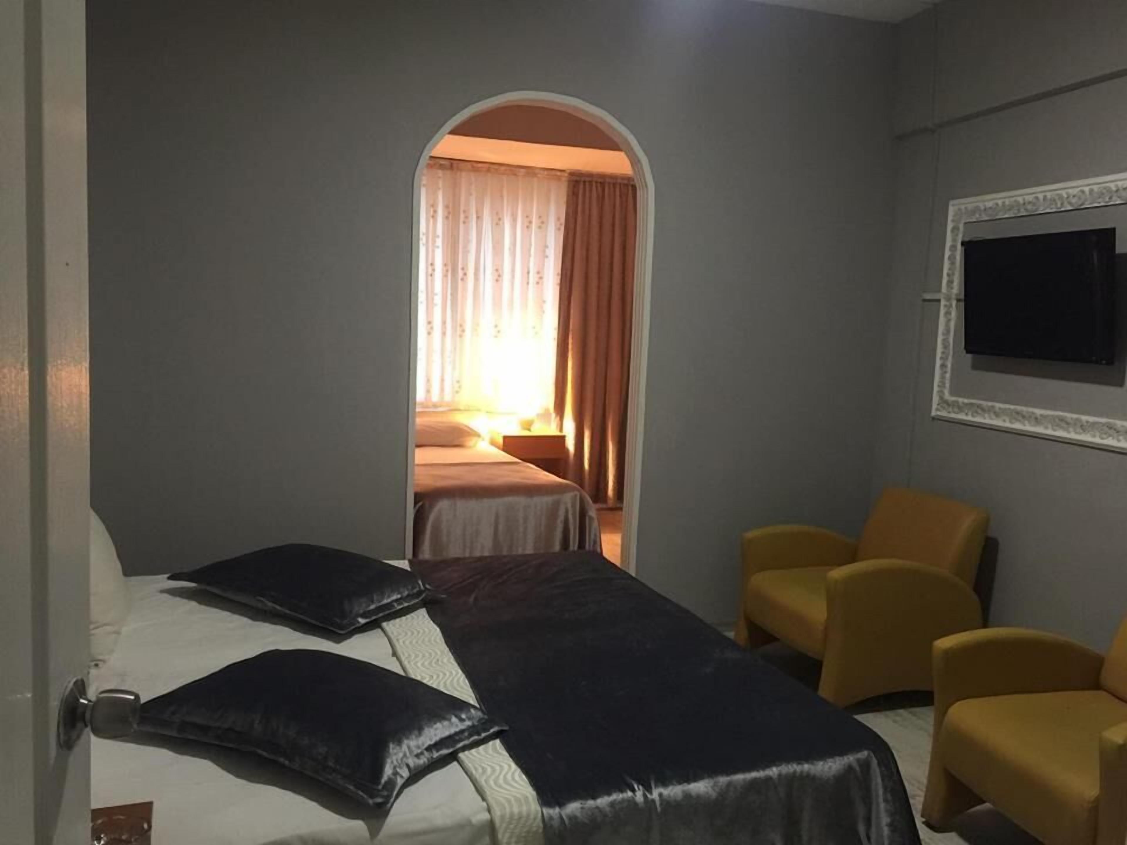 A hotel room with a bed, two yellow armchairs, a TV on the wall, and a window with curtains.