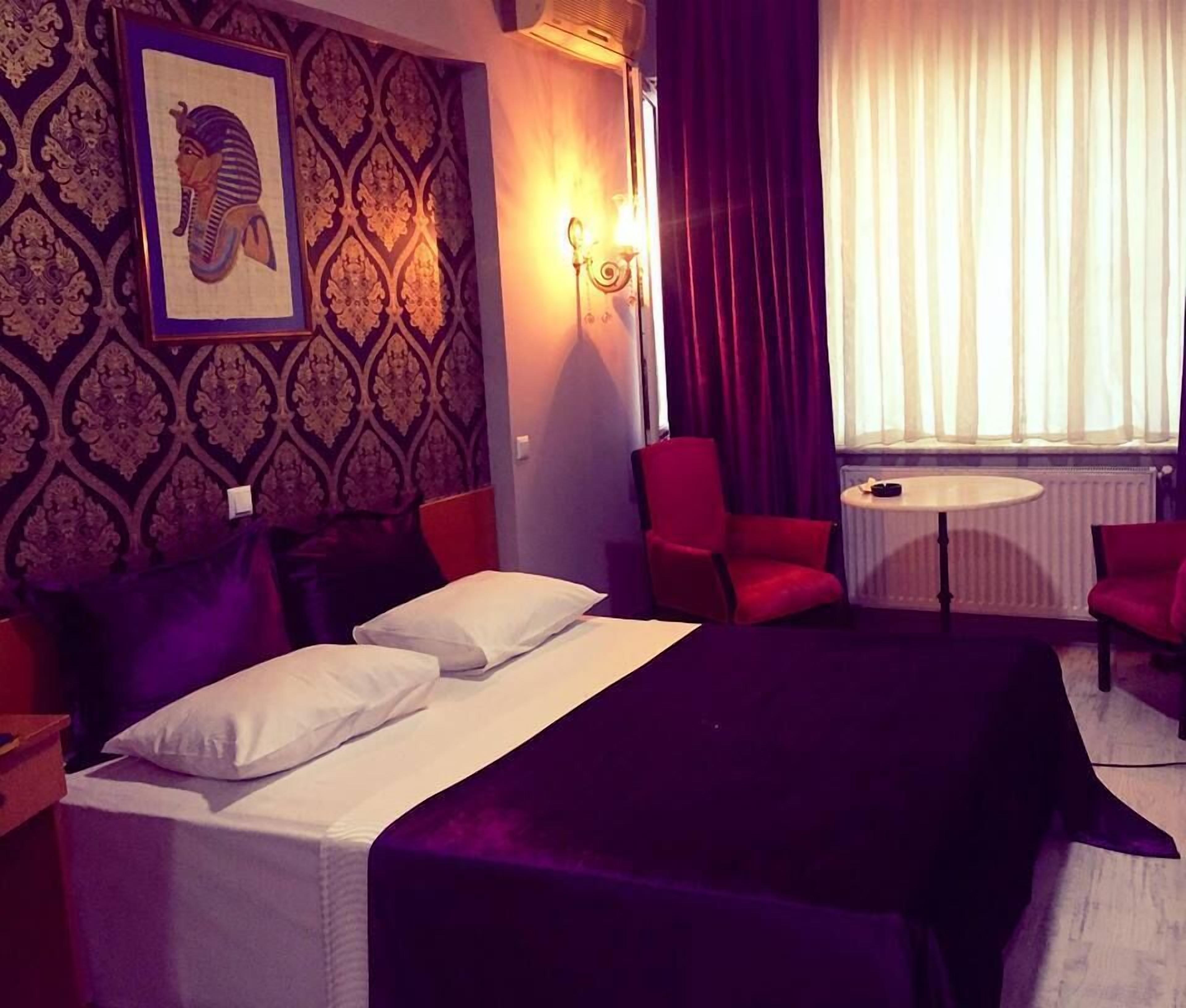 A hotel room with a bed, two red chairs, a small table, and a wall-mounted air conditioner.