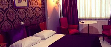 A hotel room with a bed, two red chairs, a small table, and a wall-mounted air conditioner.