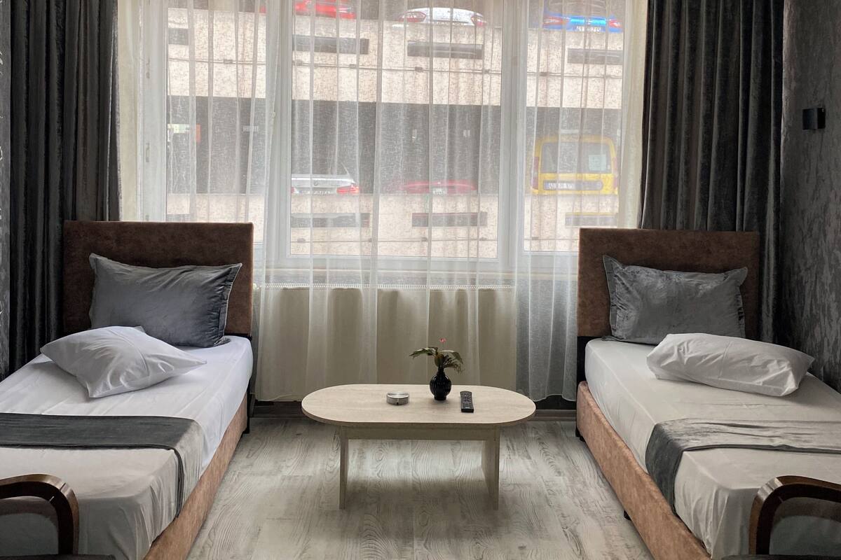 Habitación de hotel con dos camas, una mesita y vistas a la calle a través de cortinas translúcidas.