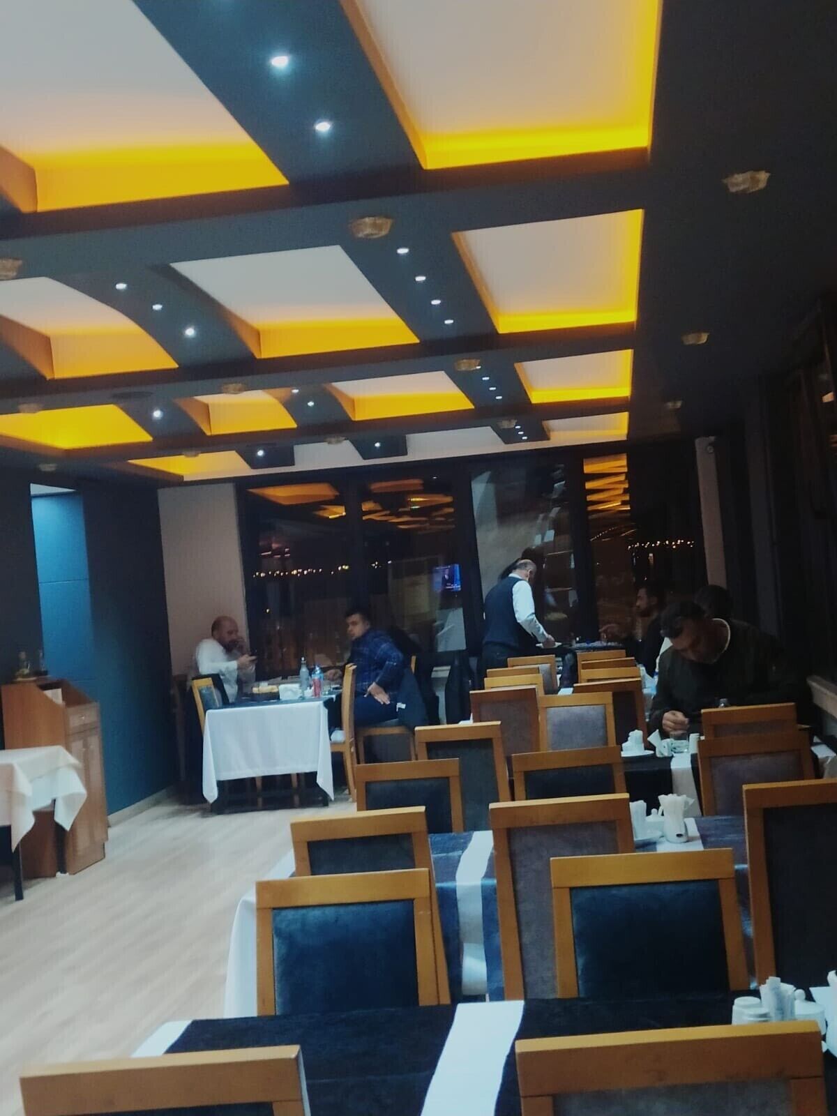 En restaurant med moderne loftdesign, der inkluderer geometriske mønstre og indbygget belysning. Borde er anbragt i en rummelig indretning med træstole.