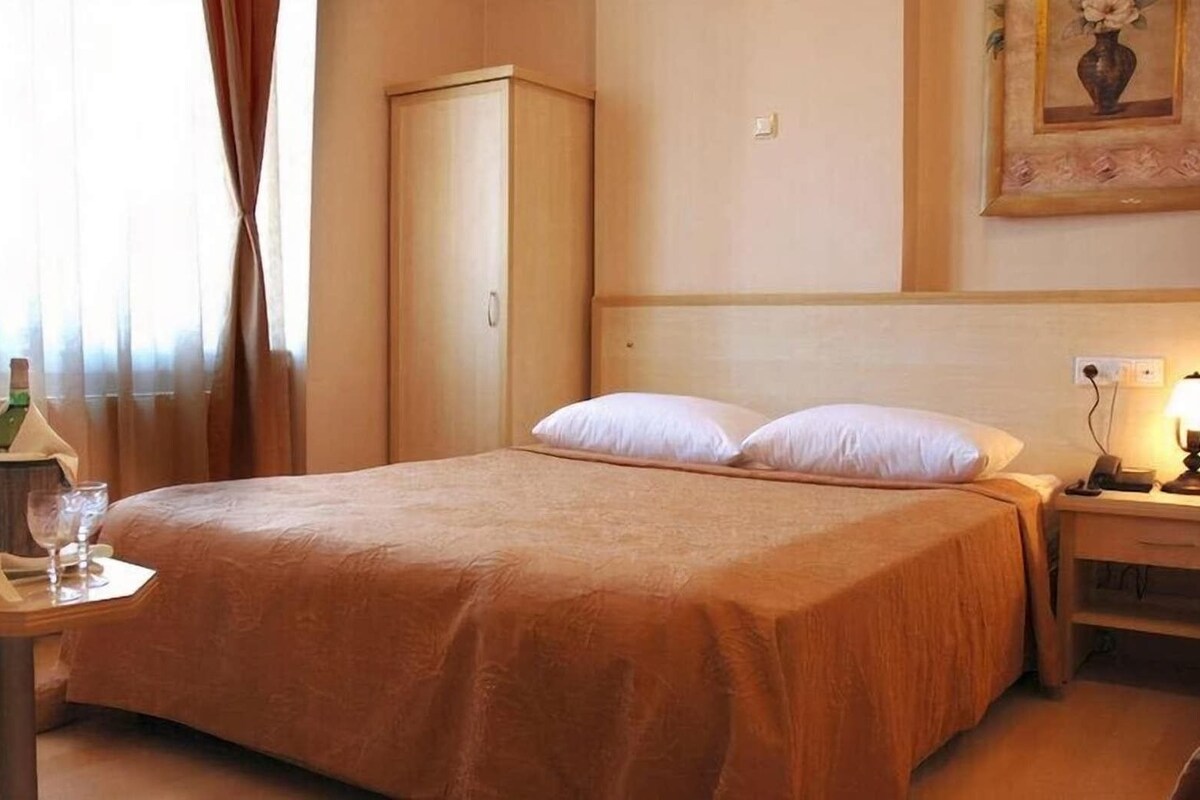 A hotel room with a bed, a bedside table with a lamp, a nightstand, a wardrobe, and a window with curtains.