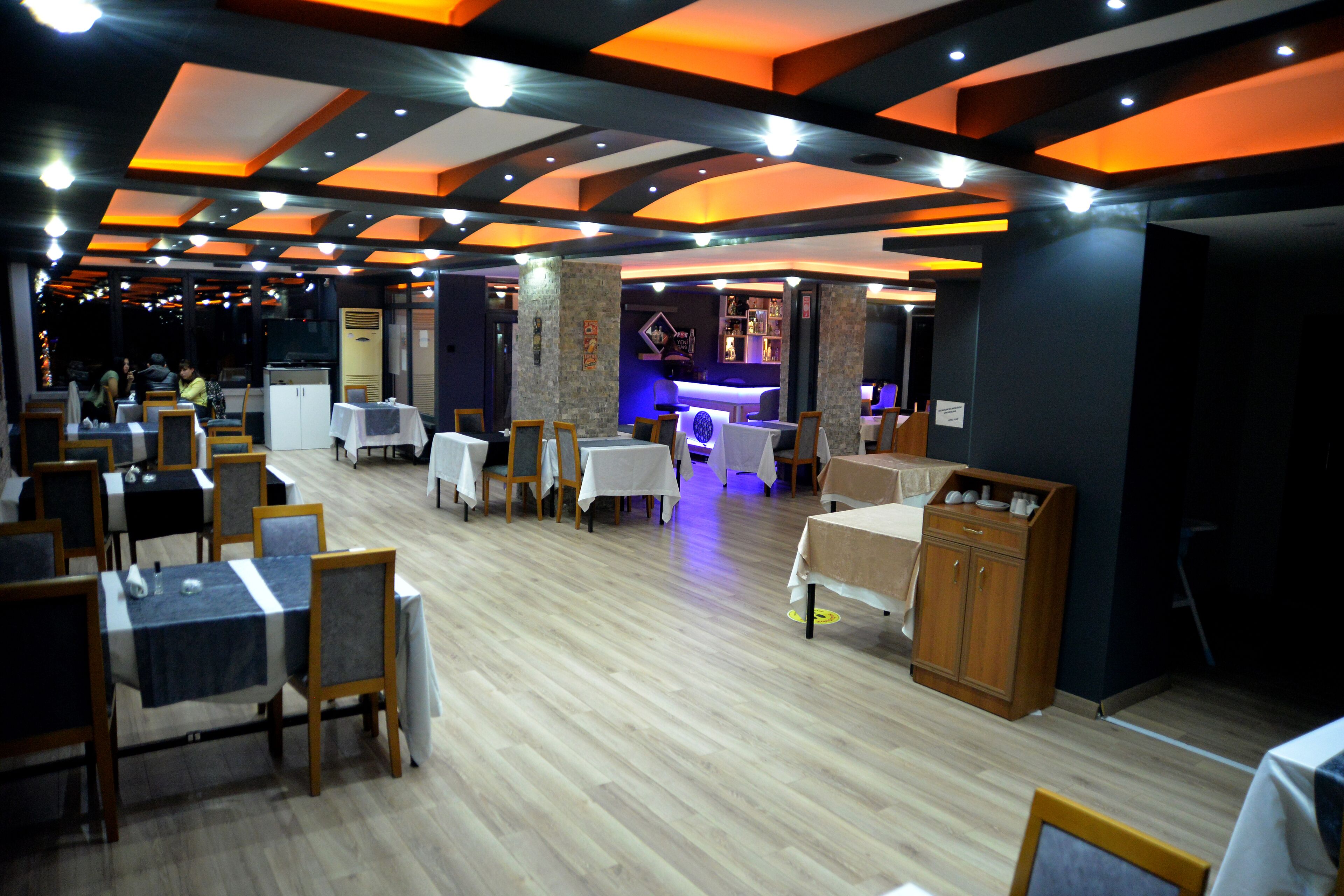 A restaurant with wooden flooring, tables with white tablecloths, and a bar area in the background.