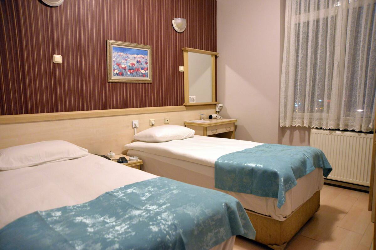 Ein Hotelzimmer mit zwei Betten, einem Nachttisch, einem Spiegel und einem Fenster mit durchsichtigen Vorhängen.