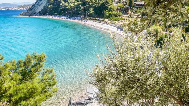 Ein Küstenstrand mit klarem türkisblauem Wasser, einem sandigen Ufer und Bäumen und Hügeln im Hintergrund.