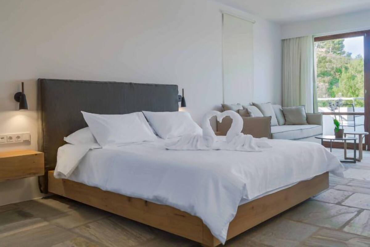 A modern bedroom with a wooden bed, white bedding, a dark headboard, a bedside table with a lamp, and a view of the outdoors through a large window.