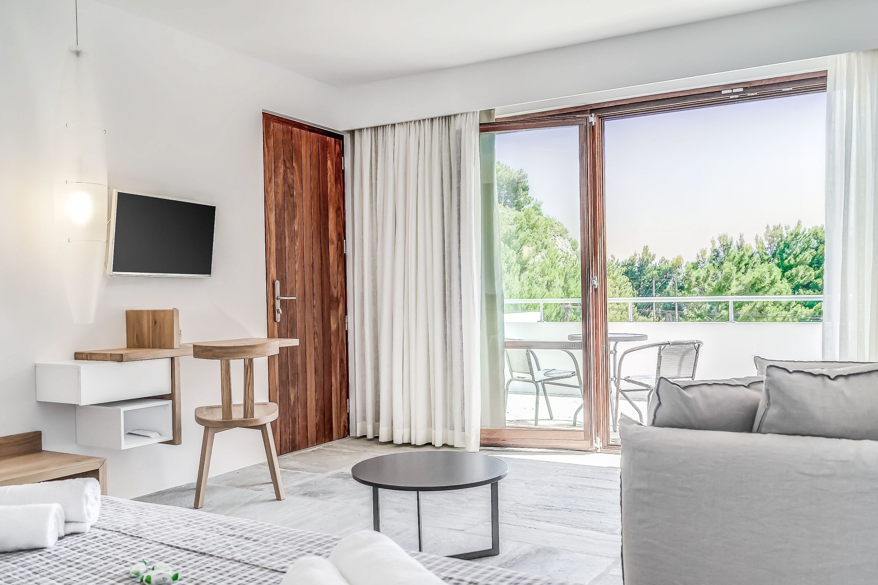 A modern living room with a sofa, a small round table, a TV mounted on the wall, and a sliding glass door leading to an outdoor area with a view of greenery.