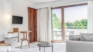 A modern living room with a sofa, a small round table, a TV mounted on the wall, and a sliding glass door leading to an outdoor area with a view of greenery.