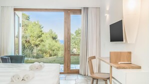 Ein Hotelzimmer mit Bett, Schreibtisch, Stuhl, Fernseher und Blick auf Grünflächen sowie ein Gebäude durch eine Glastür.