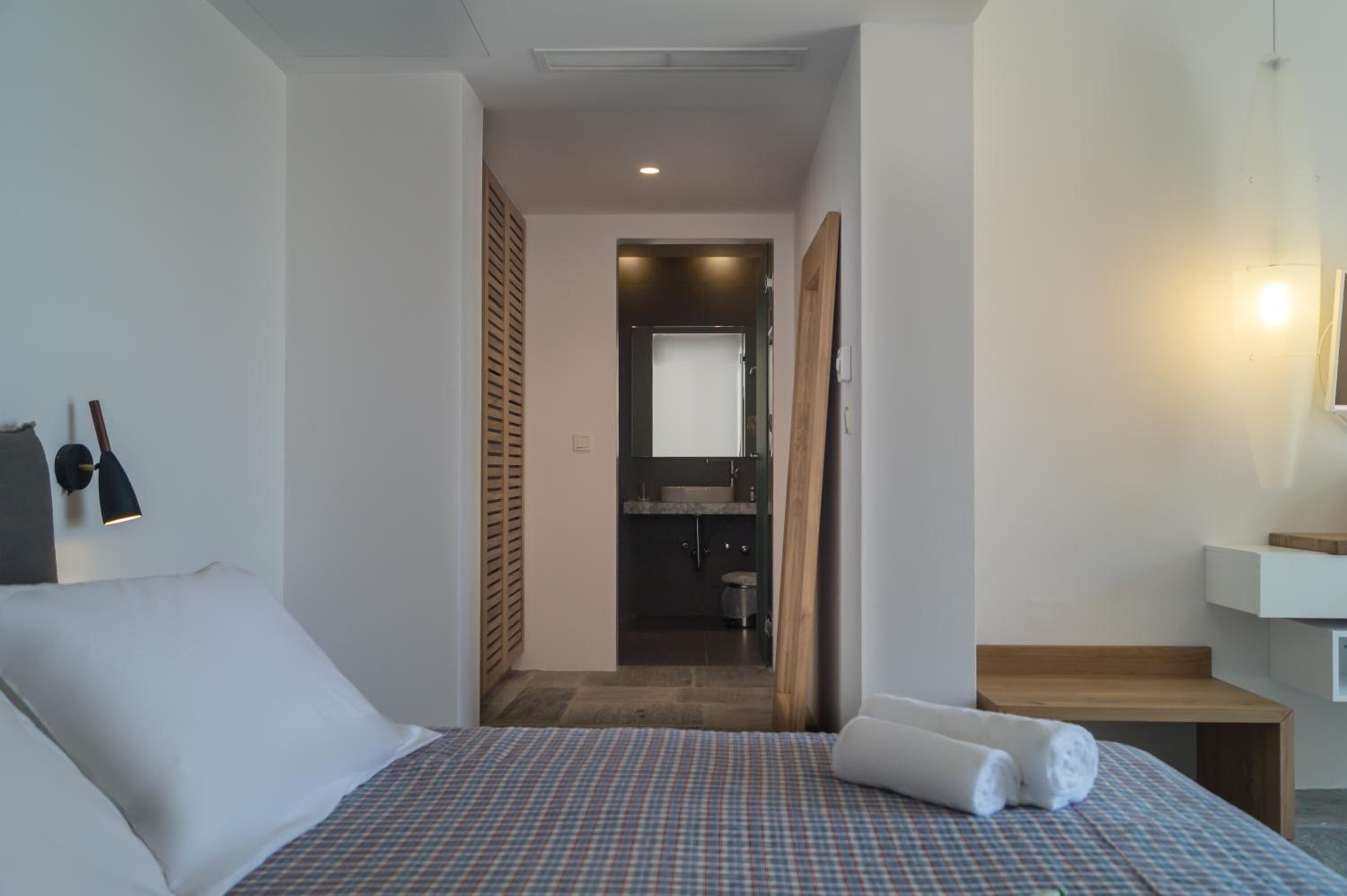 Habitación de hotel con cama, un banco de madera, una mesita, una lámpara de pared y un baño visible a través de una puerta abierta.