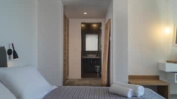 Habitación de hotel con cama, un banco de madera, una mesita, una lámpara de pared y un baño visible a través de una puerta abierta.