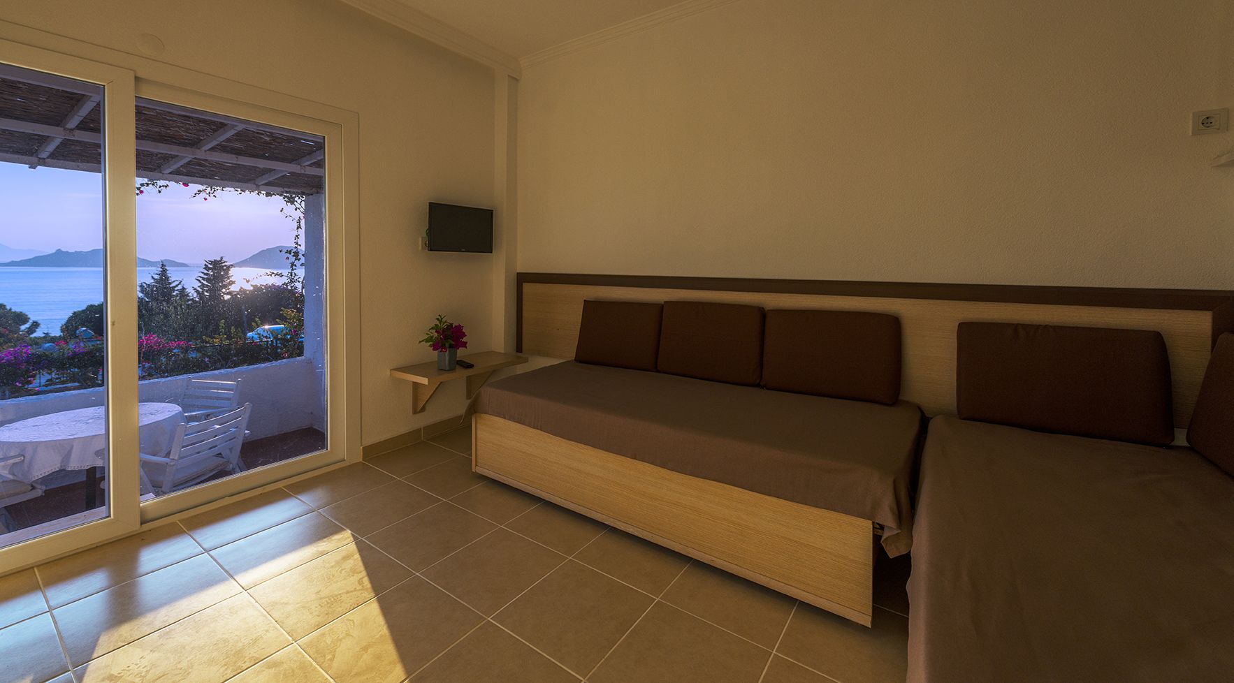 A bedroom with a bed, a nightstand, and a TV. There is a sliding glass door leading to a balcony with a table and chairs.