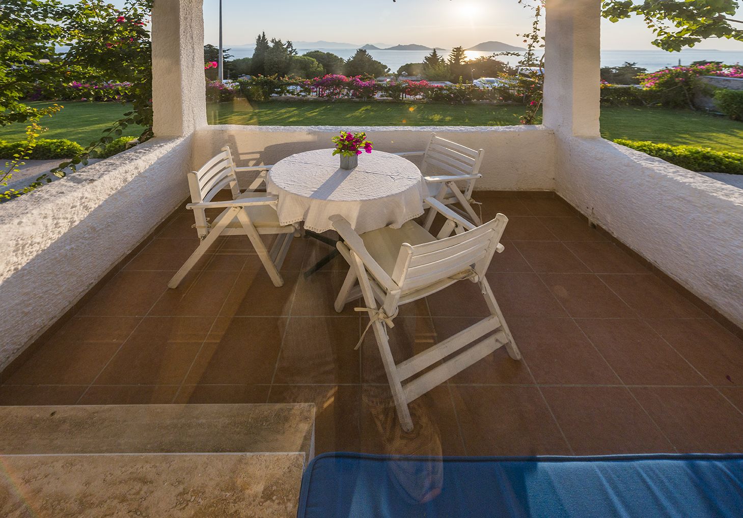 A covered outdoor patio with a round table and four chairs, a potted plant, and a view of a landscaped garden and the sea.