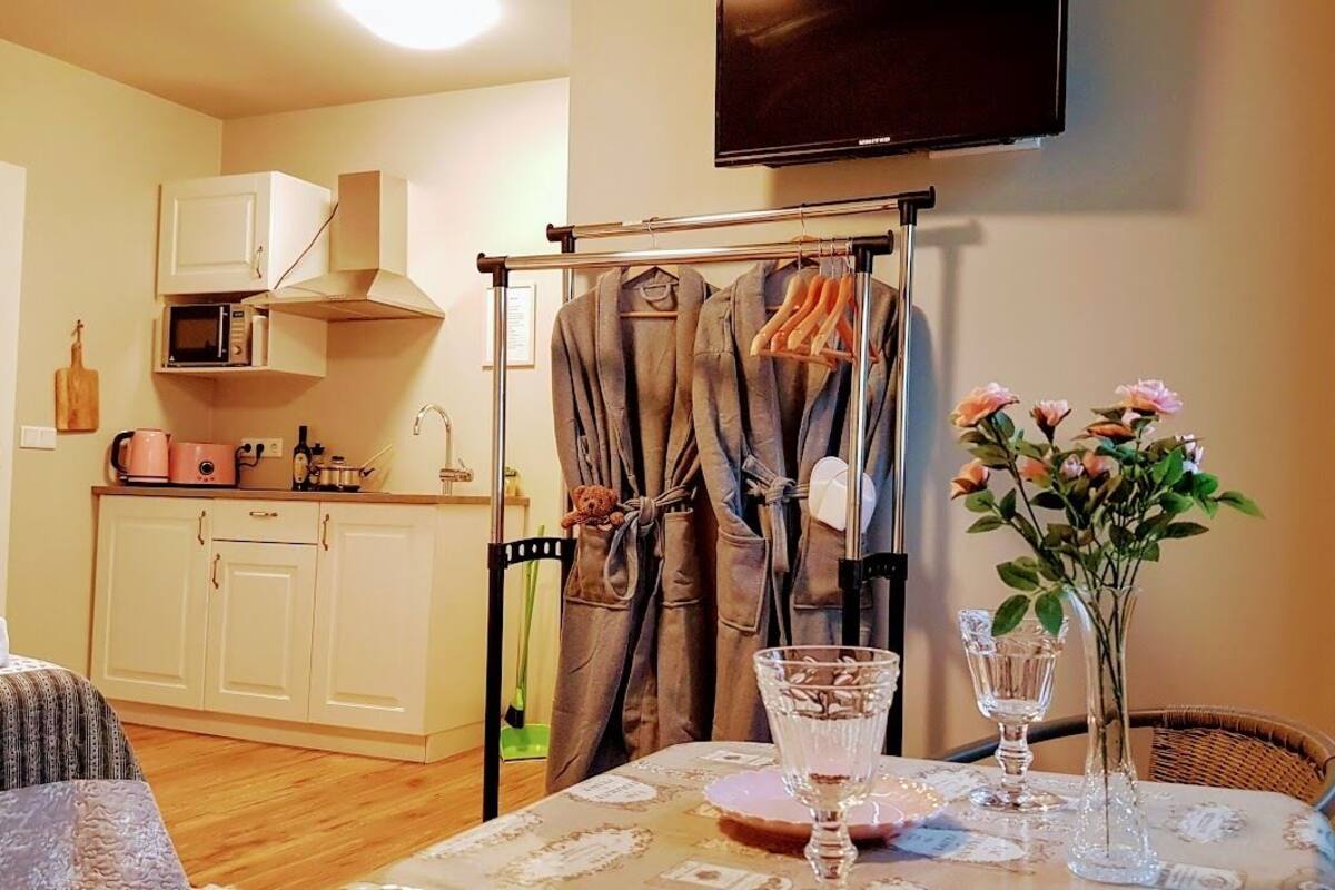 A compact kitchen with a microwave, a small sink, and cabinets. There is a dining table with a vase of flowers and two glasses.