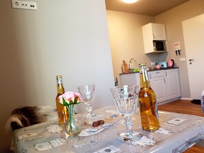 Une table à manger agrémentée d’un centre de table floral, deux bouteilles et un verre. On aperçoit une cuisine à l’arrière-plan.